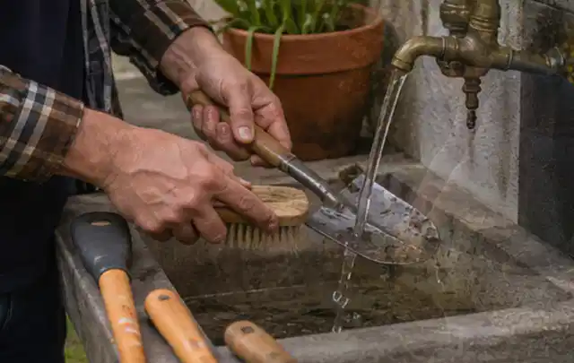Hände reinigen gebrauchte Gartengeräte mit Bürste und Wasser am Außenbecken; Thema Rasenmäher, Schwerpunkt Sicherheit; Umsetzung.
