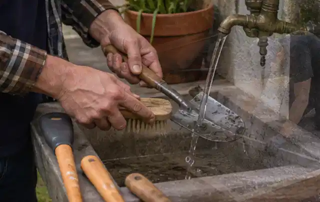 Hände reinigen gebrauchte Gartengeräte mit Bürste und Wasser am Außenbecken; Thema Rasenmäher, Schwerpunkt Systemvergleich; Ergebnis.