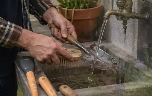 Hände reinigen gebrauchte Gartengeräte mit Bürste und Wasser am Außenbecken; Thema Rasenmäher, Schwerpunkt Wartung; Ausgangslage.