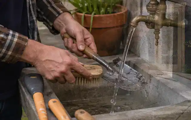 Hände reinigen gebrauchte Gartengeräte mit Bürste und Wasser am Außenbecken; Thema Rasenmäher, Schwerpunkt Winterlager; Umsetzung.