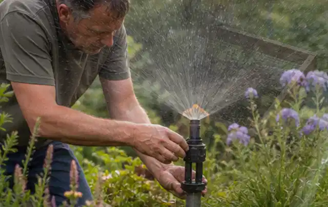 Person richtet einen laufenden Gartensprenger zwischen Stauden präzise aus; Thema Rasenunkraut, Schwerpunkt Sommerpflege; Kontrolle.