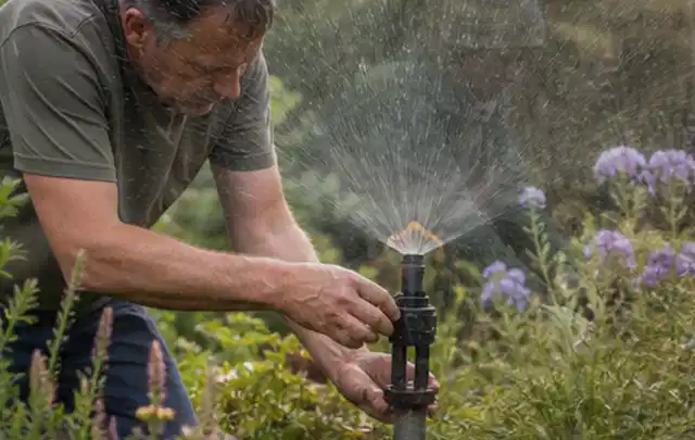 Person richtet einen laufenden Gartensprenger zwischen Stauden präzise aus; Thema Regentonnen, Schwerpunkt Sicherheit; Praxisalltag.