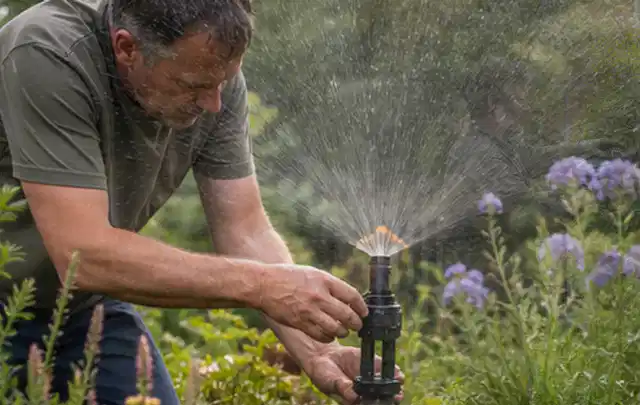 Person richtet einen laufenden Gartensprenger zwischen Stauden präzise aus; Thema Regentonnen, Schwerpunkt Wartung; Ausgangslage.