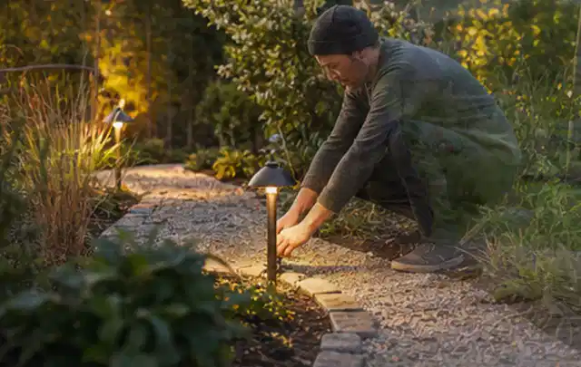 Person richtet eine niedrige warm leuchtende Gartenlampe am Kiesweg aus; Thema RTK Mähroboter, Schwerpunkt Anwendung; Ergebnis.