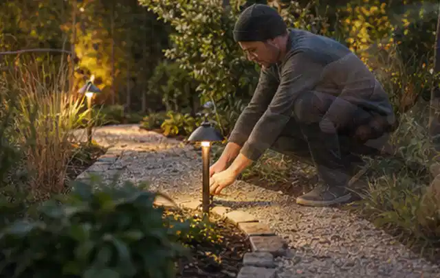 Person richtet eine niedrige warm leuchtende Gartenlampe am Kiesweg aus; Thema RTK Mähroboter, Schwerpunkt Checkliste; Umsetzung.