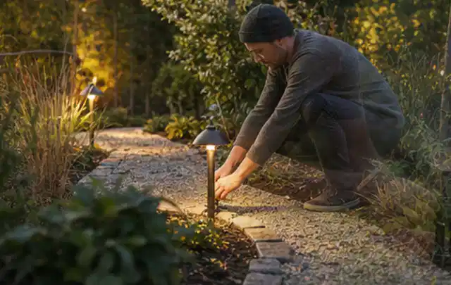 Person richtet eine niedrige warm leuchtende Gartenlampe am Kiesweg aus; Thema RTK Mähroboter, Schwerpunkt Ersatzteile; Umsetzung.