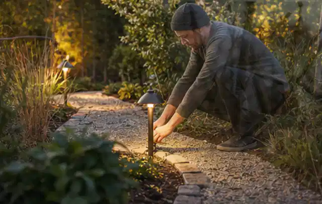 Person richtet eine niedrige warm leuchtende Gartenlampe am Kiesweg aus; Thema RTK Mähroboter, Schwerpunkt Leistung; Praxisalltag.