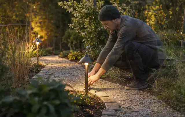 Person richtet eine niedrige warm leuchtende Gartenlampe am Kiesweg aus; Thema RTK Mähroboter, Schwerpunkt Nachhaltigkeit; Praxisalltag.