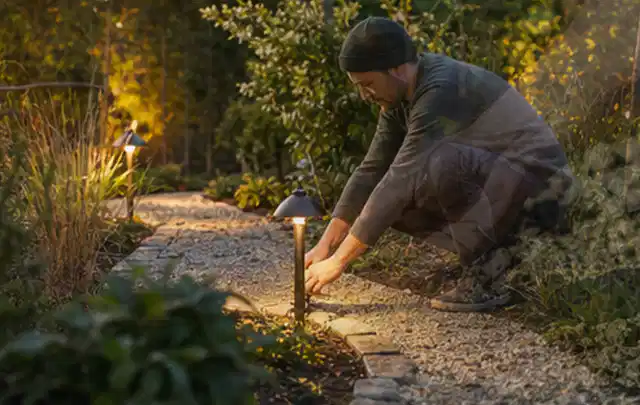 Person richtet eine niedrige warm leuchtende Gartenlampe am Kiesweg aus; Thema RTK Mähroboter, Schwerpunkt Reinigung; Ausgangslage.