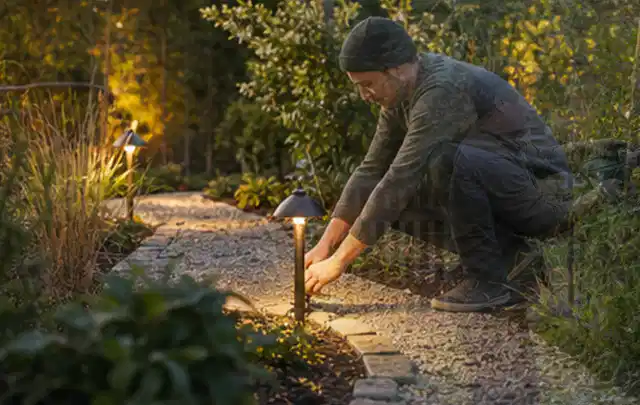 Person richtet eine niedrige warm leuchtende Gartenlampe am Kiesweg aus; Thema RTK Mähroboter, Schwerpunkt Testkriterien; Kontrolle.