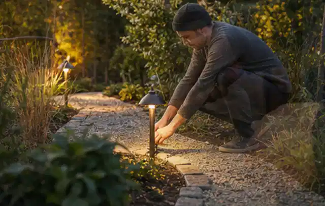 Person richtet eine niedrige warm leuchtende Gartenlampe am Kiesweg aus; Thema RTK Mähroboter, Schwerpunkt Wartung; Kontrolle.