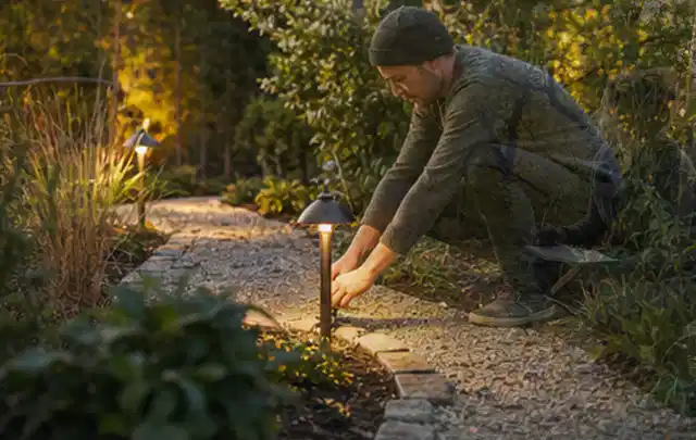 Person richtet eine niedrige warm leuchtende Gartenlampe am Kiesweg aus; Thema Salzwasserpools, Schwerpunkt Bestenliste; Kontrolle.