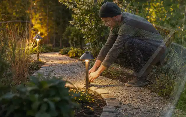 Person richtet eine niedrige warm leuchtende Gartenlampe am Kiesweg aus; Thema Salzwasserpools, Schwerpunkt Dimensionierung; Umsetzung.