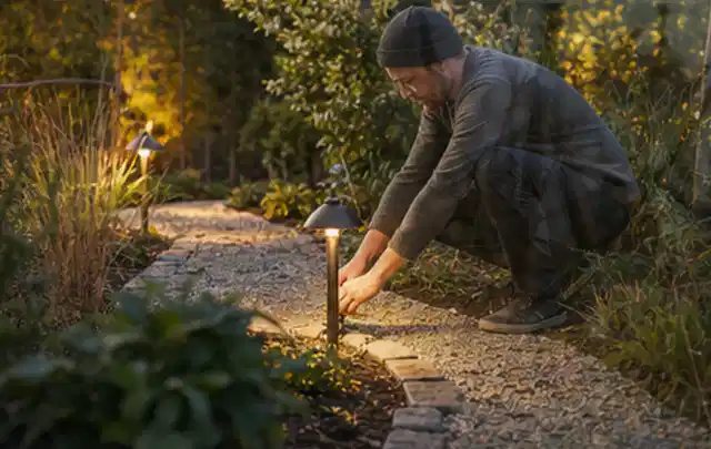 Person richtet eine niedrige warm leuchtende Gartenlampe am Kiesweg aus; Thema Salzwasserpools, Schwerpunkt Leistung; Kontrolle.