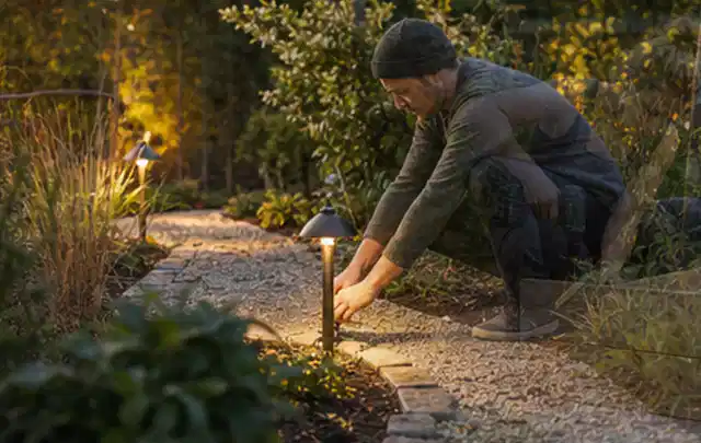 Person richtet eine niedrige warm leuchtende Gartenlampe am Kiesweg aus; Thema Salzwasserpools, Schwerpunkt Nachhaltigkeit; Kontrolle.