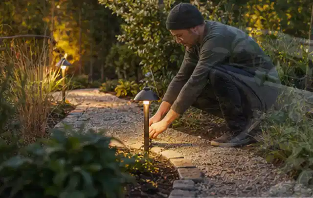 Person richtet eine niedrige warm leuchtende Gartenlampe am Kiesweg aus; Thema Salzwasserpools, Schwerpunkt Sicherheit; Umsetzung.
