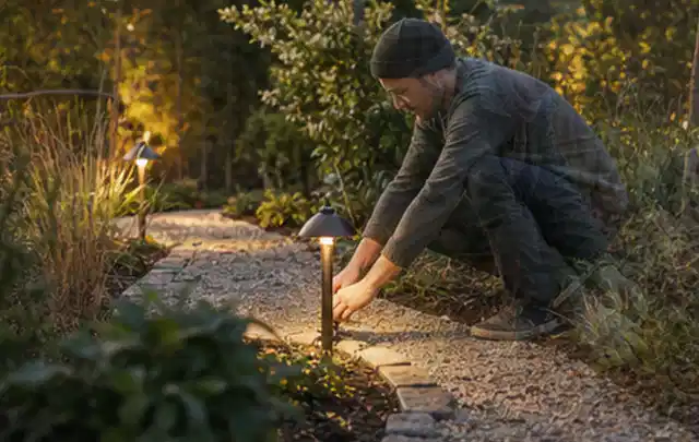 Person richtet eine niedrige warm leuchtende Gartenlampe am Kiesweg aus; Thema Salzwasserpools, Schwerpunkt Systemvergleich; Praxisalltag.