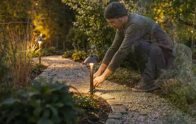 Person richtet eine niedrige warm leuchtende Gartenlampe am Kiesweg aus; Thema Salzwasserpools, Schwerpunkt Wartung; Ergebnis.