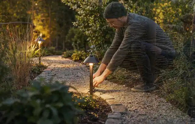 Person richtet eine niedrige warm leuchtende Gartenlampe am Kiesweg aus; Thema Sandfilteranlagen, Schwerpunkt Anwendung; Ergebnis.