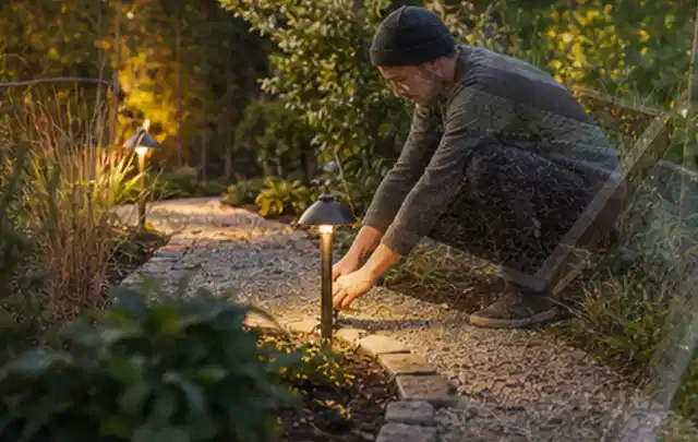 Person richtet eine niedrige warm leuchtende Gartenlampe am Kiesweg aus; Thema Sandfilteranlagen, Schwerpunkt Dimensionierung; Umsetzung.