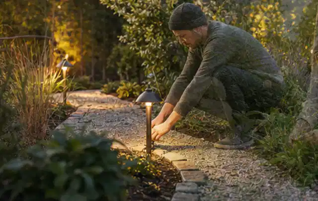Person richtet eine niedrige warm leuchtende Gartenlampe am Kiesweg aus; Thema Sandfilteranlagen, Schwerpunkt Nachhaltigkeit; Ausgangslage.