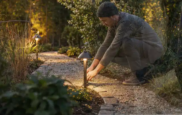 Person richtet eine niedrige warm leuchtende Gartenlampe am Kiesweg aus; Thema Sandfilteranlagen, Schwerpunkt Reinigung; Umsetzung.