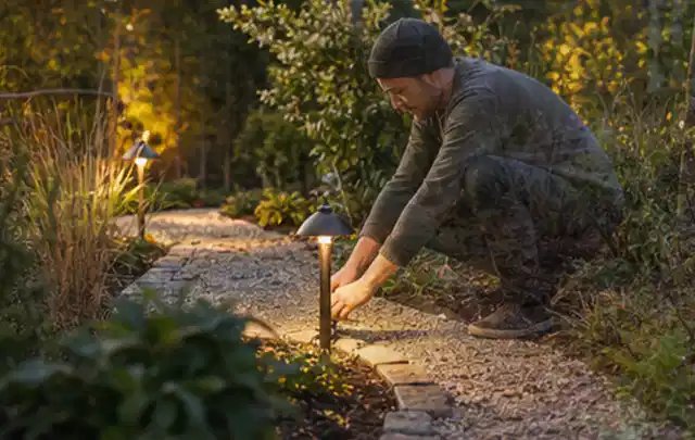 Person richtet eine niedrige warm leuchtende Gartenlampe am Kiesweg aus; Thema Sandfilteranlagen, Schwerpunkt Systemvergleich; Ausgangslage.
