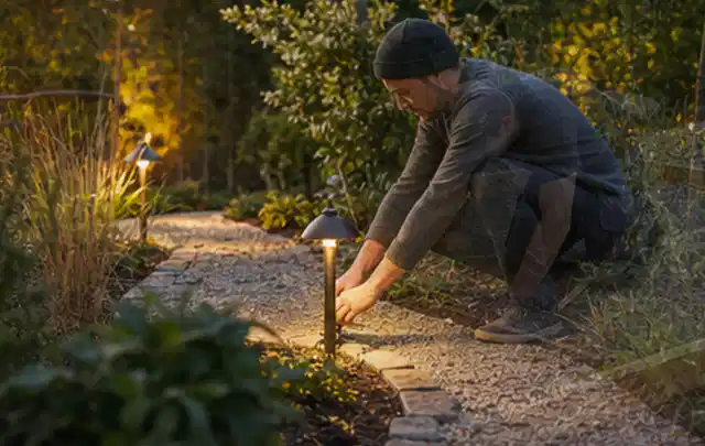 Person richtet eine niedrige warm leuchtende Gartenlampe am Kiesweg aus; Thema Sandfilteranlagen, Schwerpunkt Testkriterien; Umsetzung.
