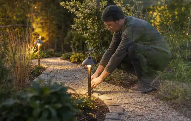 Person richtet eine niedrige warm leuchtende Gartenlampe am Kiesweg aus; Thema Segway Navimow, Schwerpunkt Checkliste; Umsetzung.