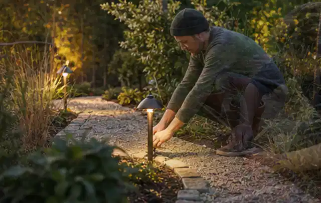 Person richtet eine niedrige warm leuchtende Gartenlampe am Kiesweg aus; Thema Segway Navimow, Schwerpunkt Systemvergleich; Ausgangslage.