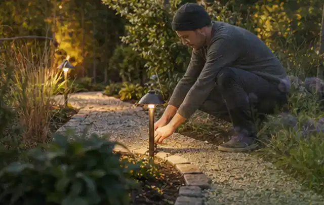 Person richtet eine niedrige warm leuchtende Gartenlampe am Kiesweg aus; Thema Segway Navimow, Schwerpunkt Vergleich; Ergebnis.