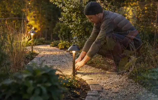 Person richtet eine niedrige warm leuchtende Gartenlampe am Kiesweg aus; Thema Sichtschutz aus Holz, Schwerpunkt Aufbau; Umsetzung.
