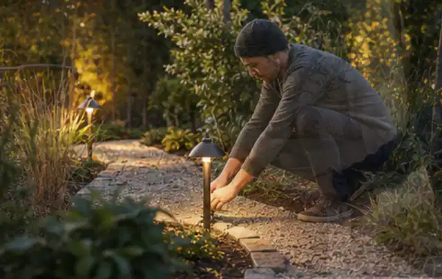 Person richtet eine niedrige warm leuchtende Gartenlampe am Kiesweg aus; Thema Sichtschutz aus Holz, Schwerpunkt Baurecht; Umsetzung.
