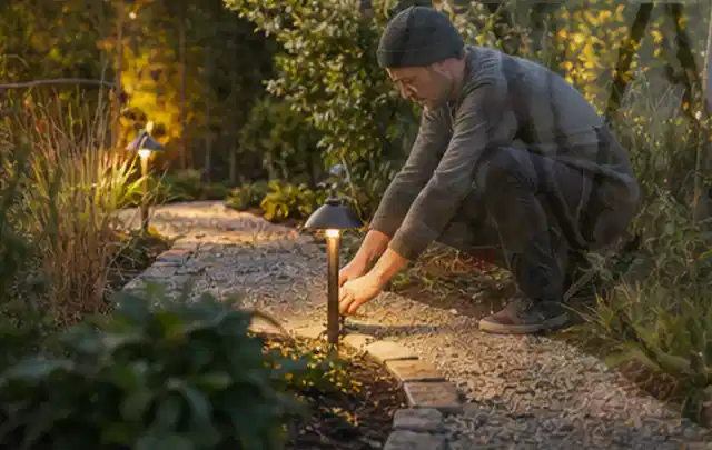 Person richtet eine niedrige warm leuchtende Gartenlampe am Kiesweg aus; Thema Sichtschutz aus Holz, Schwerpunkt DIY; Kontrolle.