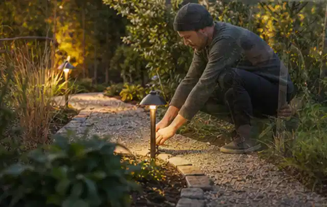 Person richtet eine niedrige warm leuchtende Gartenlampe am Kiesweg aus; Thema Sichtschutz aus Holz, Schwerpunkt Gestaltung; Ausgangslage.