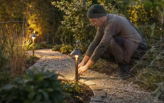 Person richtet eine niedrige warm leuchtende Gartenlampe am Kiesweg aus; Thema Sichtschutz aus Holz, Schwerpunkt Nachhaltigkeit; Kontrolle.