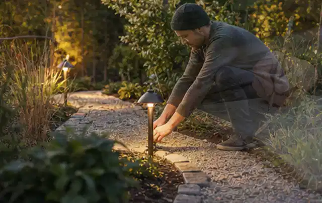 Person richtet eine niedrige warm leuchtende Gartenlampe am Kiesweg aus; Thema Sichtschutzhecken, Schwerpunkt Baurecht; Ergebnis.