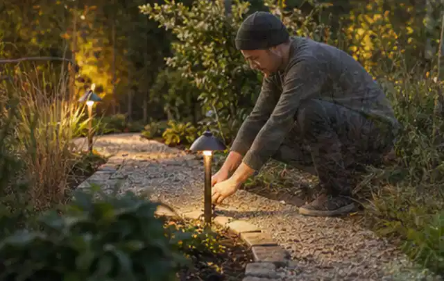Person richtet eine niedrige warm leuchtende Gartenlampe am Kiesweg aus; Thema Sichtschutzhecken, Schwerpunkt Gärten mit kleiner Fläche; Ergebnis.