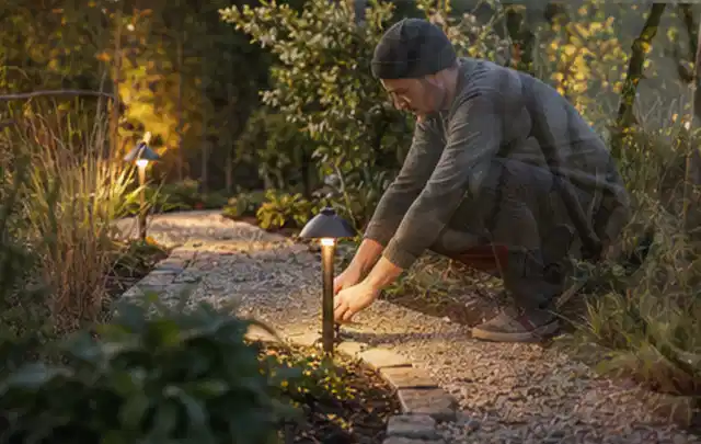 Person richtet eine niedrige warm leuchtende Gartenlampe am Kiesweg aus; Thema Sichtschutzhecken, Schwerpunkt Gestaltung; Umsetzung.
