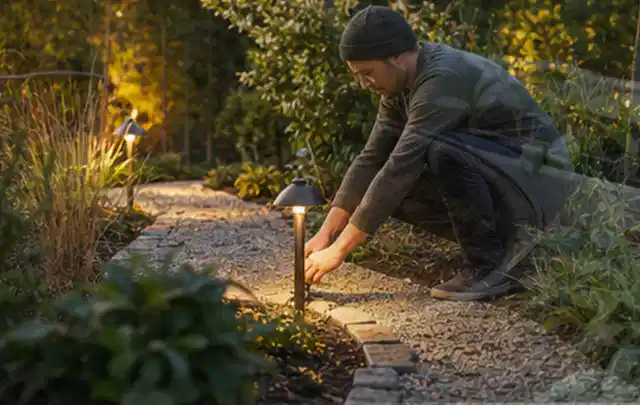 Person richtet eine niedrige warm leuchtende Gartenlampe am Kiesweg aus; Thema Sichtschutzhecken, Schwerpunkt Nachhaltigkeit; Ergebnis.