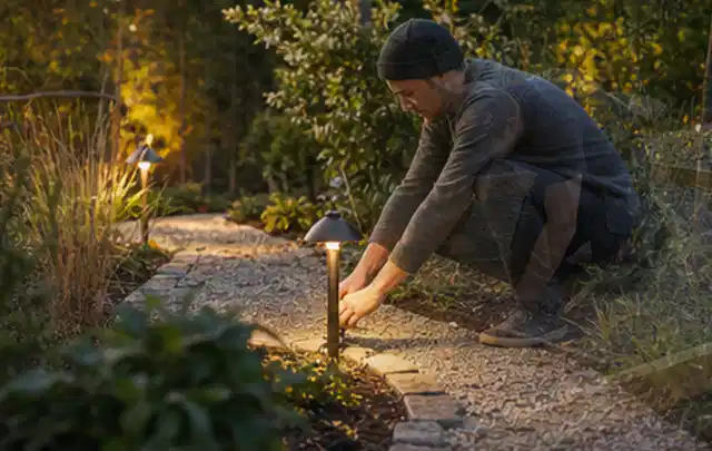 Person richtet eine niedrige warm leuchtende Gartenlampe am Kiesweg aus; Thema Sichtschutzmatten, Schwerpunkt Aufbau; Kontrolle.