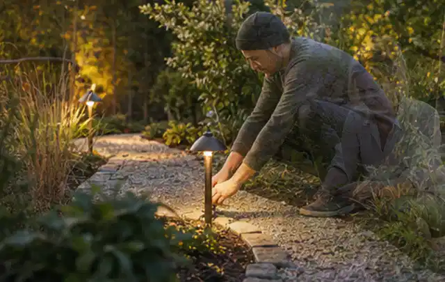 Person richtet eine niedrige warm leuchtende Gartenlampe am Kiesweg aus; Thema Sichtschutzmatten, Schwerpunkt Dimensionierung; Kontrolle.