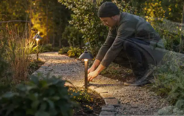 Person richtet eine niedrige warm leuchtende Gartenlampe am Kiesweg aus; Thema Sichtschutzmatten, Schwerpunkt Fundament; Umsetzung.