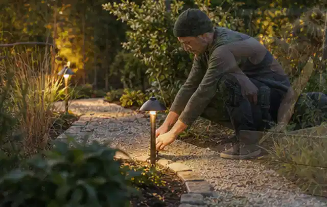 Person richtet eine niedrige warm leuchtende Gartenlampe am Kiesweg aus; Thema Sichtschutzmatten, Schwerpunkt Materialwahl; Ausgangslage.