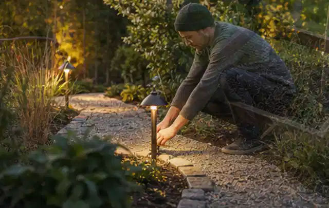 Person richtet eine niedrige warm leuchtende Gartenlampe am Kiesweg aus; Thema Sichtschutzmatten, Schwerpunkt Pflege; Praxisalltag.