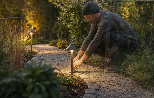 Person richtet eine niedrige warm leuchtende Gartenlampe am Kiesweg aus; Thema Sichtschutzmatten, Schwerpunkt Planung; Ergebnis.