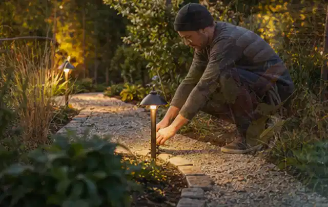 Person richtet eine niedrige warm leuchtende Gartenlampe am Kiesweg aus; Thema Sichtschutzmatten, Schwerpunkt Winterschutz; Umsetzung.