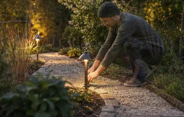 Person richtet eine niedrige warm leuchtende Gartenlampe am Kiesweg aus; Thema Smoker, Schwerpunkt Dimensionierung; Praxisalltag.