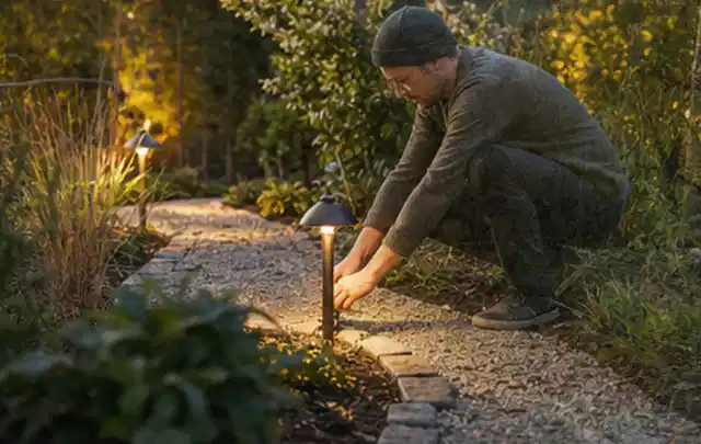 Person richtet eine niedrige warm leuchtende Gartenlampe am Kiesweg aus; Thema Smoker, Schwerpunkt Gärten mit großer Fläche; Praxisalltag.