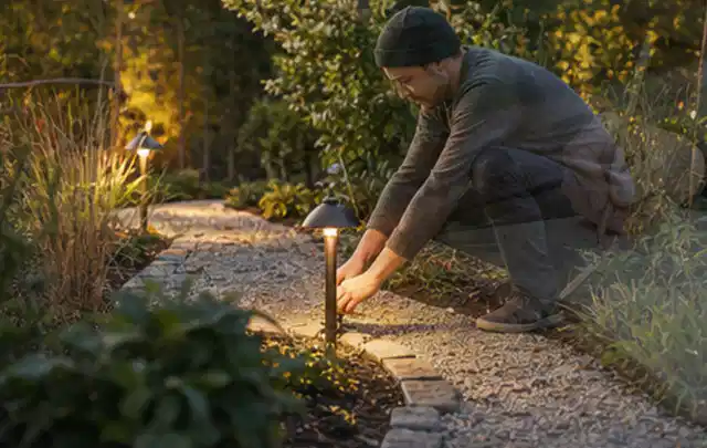 Person richtet eine niedrige warm leuchtende Gartenlampe am Kiesweg aus; Thema Smoker, Schwerpunkt Reinigung; Umsetzung.